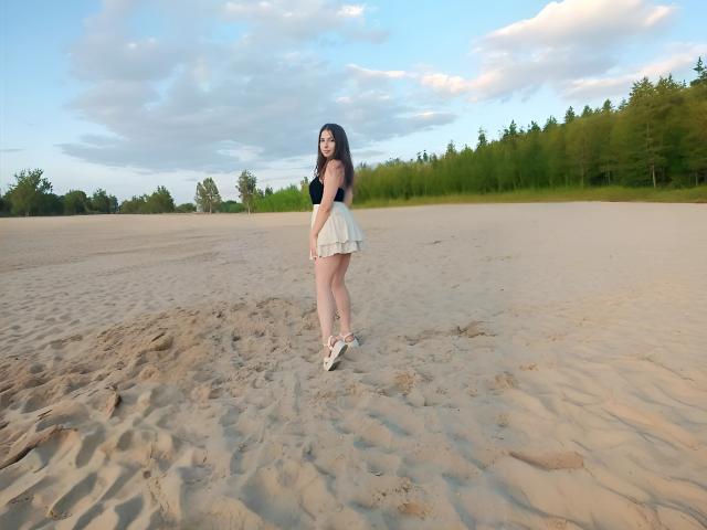DominikaRos met lang donker haar, kleine borsten, een witte rok met ruches, een zwarte top, staand op een zandstrand, kijkend achterom, handen achter de rug, witte schoenen, een groen bos op de achtergrond..