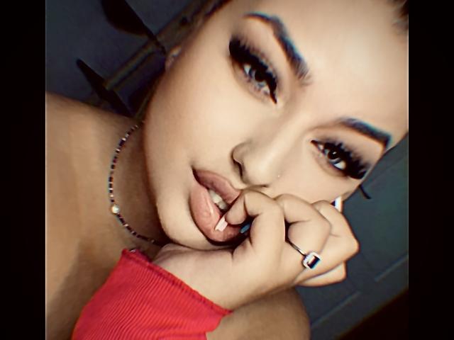 AllysonK with blue eyebrows, long eyelashes, and a nose ring, biting her finger with red shirt, silver necklace, and blue ring, looking at camera with sexy expression.