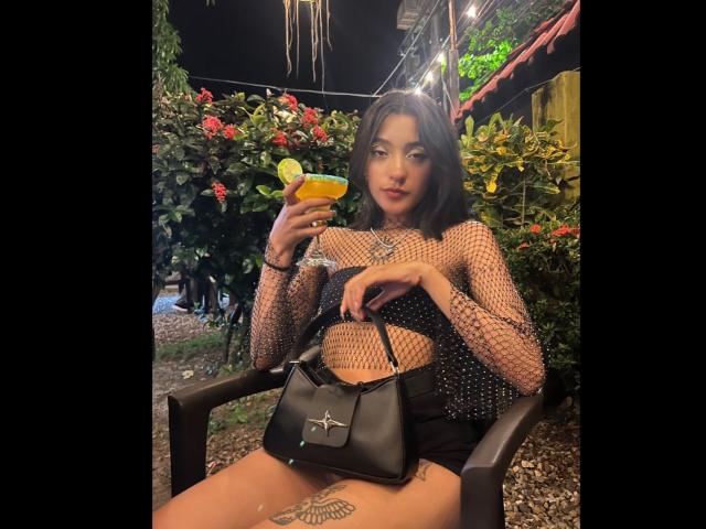 SalomeMille, met donker haar, draagt een top van netstof, zwarte shorts en een zwarte tas, ze houdt een cocktail met een citroenschijfje vast, zit op een stoel en toont haar getatoeëerde vulva..