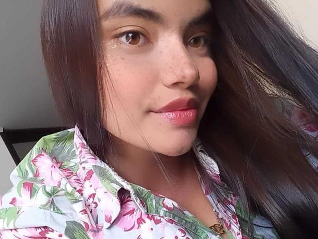 BeckyKx with long brown hair, brown eyes, pink glossy lips, wearing a floral shirt, slight smile, light freckles on her nose, soft skin, casual selfie.