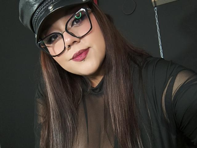 EmilyDom with long brown hair, black glasses, and a black leather cap, wearing a sheer black top, showing a pierced lip and a subtle smile, looking at the camera.