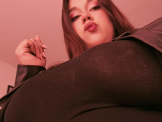 EmilyDom con labios rojos y gruesos, cabello oscuro y ondulado, una blusa negra texturizada y una chaqueta de cuero negro, mostrando sus grandes senos en una toma desde un ángulo bajo..