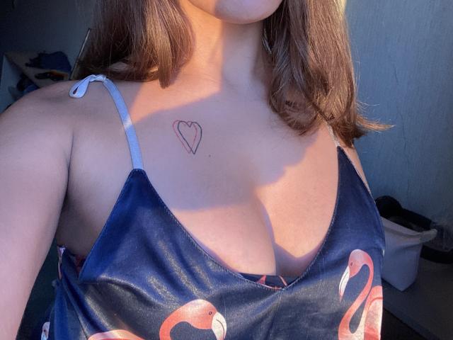 Os seios de Kametia estão visíveis em um vestido azul com estampa de flamingos rosa, com uma pequena tatuagem de coração acima de sua região do colo, em uma selfie parcial tirada sob a luz do sol..