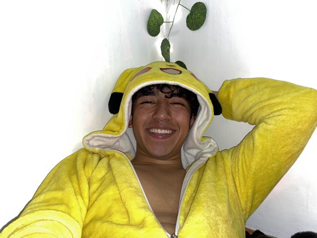 AresMc vistiendo una sudadera amarilla de Pikachu, sonriendo con los ojos cerrados, con el brazo derecho detrás de la cabeza, mostrando el pecho desnudo, cabello castaño rizado y un fondo de pared blanca con una planta verde..