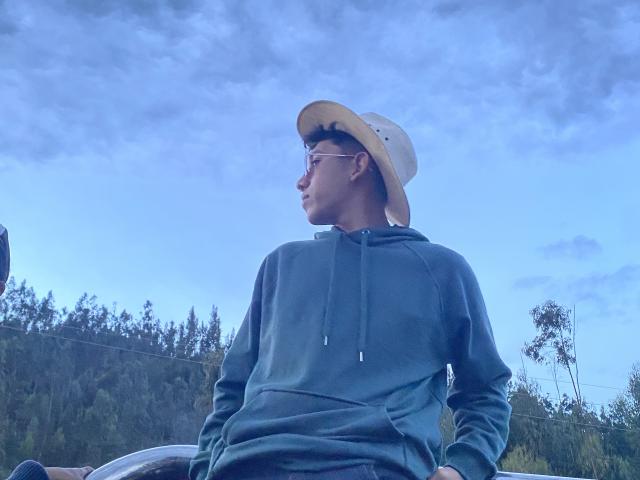 AresMc, con un sombrero beige y gafas, vistiendo una sudadera verde, con las manos en los bolsillos, de pie contra un cielo azul, mirando hacia un lado, con un bosque de fondo..