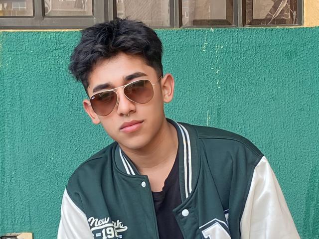 AresMc con cabello negro corto, gafas de sol estilo aviador marrones, chaqueta deportiva verde y blanca, camisa negra y expresión neutra, de pie frente a una pared verde con textura..