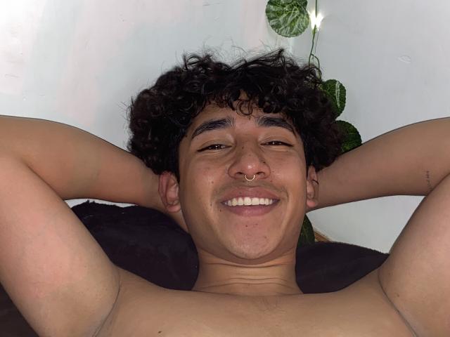 AresMc with curly dark hair, nose ring, and smiling, shirtless, arms behind head, showing a smooth, tan, slightly hairy chest, and a relaxed, happy expression.