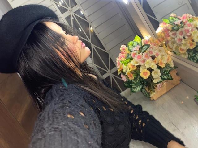 MiiaPalmer with black hat, long black hair, and black crochet top, taking selfie, showing off her tits, pink lips, and pinkish nose, next to a flower bouquet mirror.