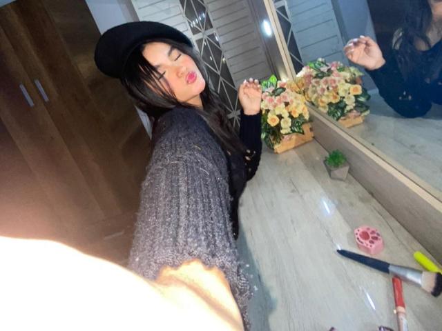 MiiaPalmer with black hat, long brown hair, and black sweater, taking selfie with raised arm, showing reflection in mirror, gray wooden floor, pink nail tool, yellow brush.