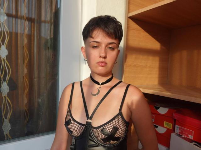 MistressMaeveFirst with short black hair, a crescent moon necklace, and a black fishnet top showing her small tits and black nipples. Standing near wooden shelves and a window.