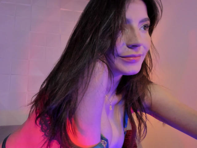 ValenRosee with long brown hair, smiling, wearing a lace bra, showcasing her boobs, purple and pink lighting, necklace, leaning forward, soft expression, casual, sexy, relaxed.