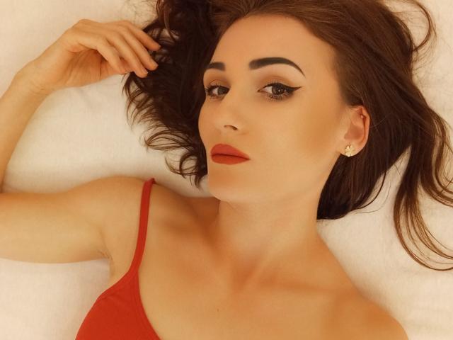 Anyy has red lipstick, dark eyebrows, and brown hair, lying on a white surface, wearing a red top, with a gold earring, and a slight smirk on her face.