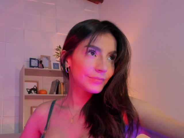 ValenRosee met roze en paarse verlichting op haar gezicht, lang bruin haar, kleine zwarte oorbellen, een ketting en een groene tanktop, met een lichte glimlach, waarbij haar borsten zichtbaar zijn..
