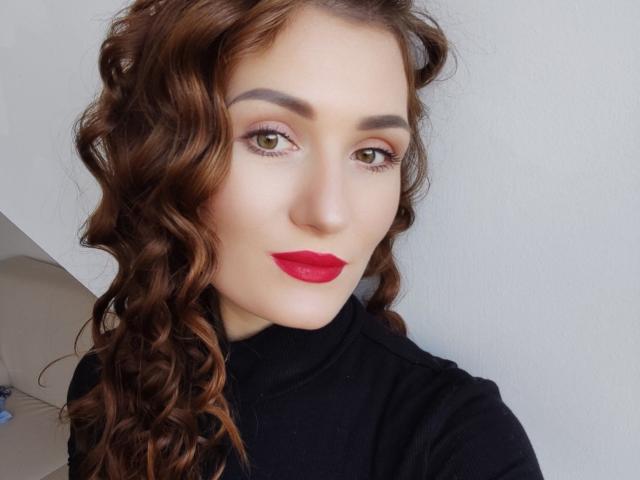 A Anny tem cabelo castanho encaracolado, olhos verdes e batom vermelho. Ela está vestindo uma gola alta preta e tem um sorriso discreto. Suas sobrancelhas são grossas e bem definidas..