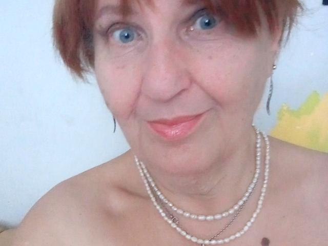 MioritaStar with short red hair, blue eyes, and pink lipstick, wearing a pearl necklace, showing bare shoulders, smiling slightly. Her tits are partially visible.