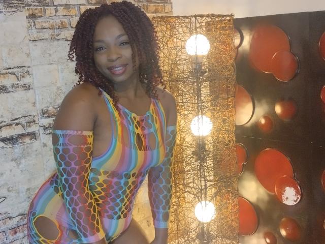 SwettBrownn, con piel oscura, cabello rizado y rojo, vestida con un traje de baño entero de manga larga, colorido y con encaje, que deja ver sus pechos a través del diseño, sonriendo a la cámara..