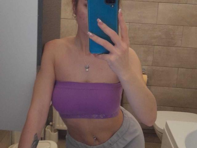 IngridI lleva un top morado que deja ver sus pechos, pantalones grises y un piercing en el ombligo, y está tomando una foto de sí misma frente a un espejo con un teléfono azul en un baño..