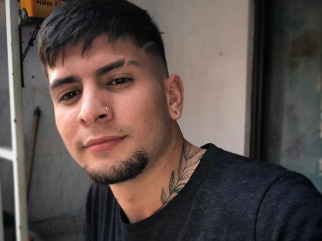 Sthefano has short black hair, a trimmed mustache, a goatee, a nose ring, and a tattoo on his neck. He's wearing a black shirt, looking slightly to the side.