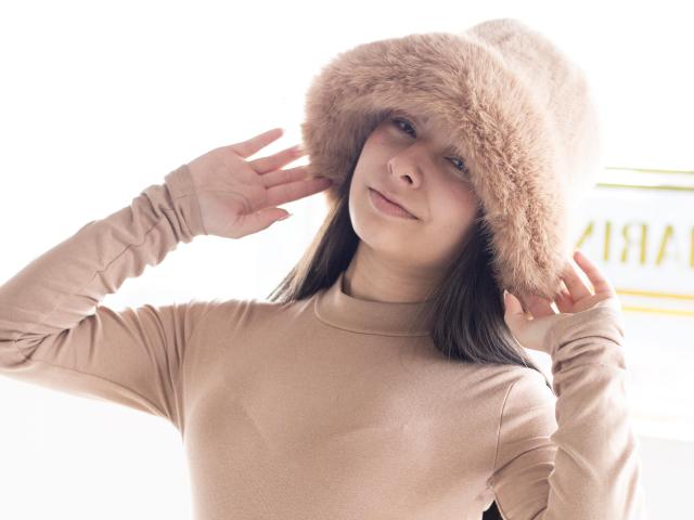 Abbie69 wearing a fluffy pinkish-brown fur hat, light beige long-sleeve top, touching hat with both hands, smiling softly, light skin, brown hair, cute, casual winter outfit.