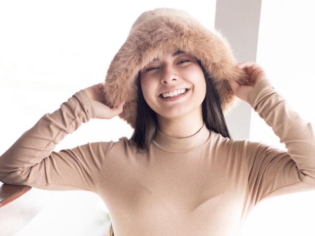 Abbie69 com um chapéu de pele marrom, sorrindo, com as mãos no chapéu, vestindo uma blusa bege de manga comprida, mostrando dentes bonitos, seios médios, pele clara, cabelo preto liso, luz interna forte..