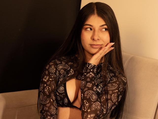 Abbie69, con lunghi capelli scuri, indossa una maglia nera con un motivo in pizzo che rivela il suo seno piccolo, appoggiando il mento sulla mano e guardando di lato con un sorriso appena accennato..