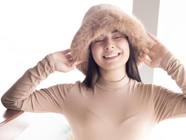 Abbie69 with eyes closed, smiling, wearing a beige furry hat and matching long-sleeve top, hands adjusting the hat, showing her cute face and small tits.