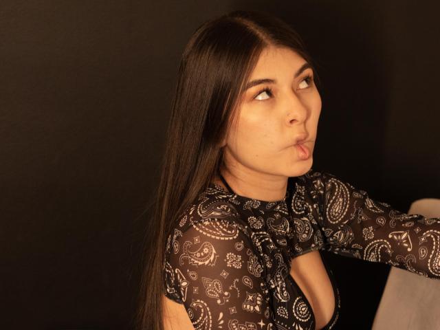 Abbie69 heeft lang zwart haar, draagt een zwarte, doorschijnende top met een diep decolleté, zet haar lippen op, kijkt naar de zijkant, op een warme, donkere achtergrond..