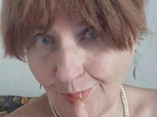 MioritaStar with short brown hair, blue eyes, and a pearl necklace, eating a finger with a small piece of food on her lips. Her expression is slightly smug.