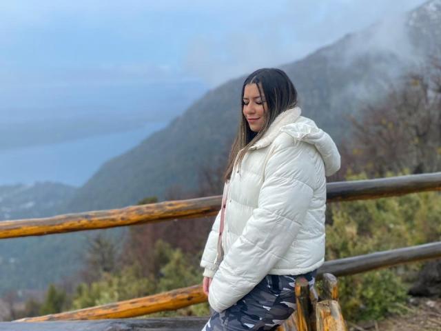 AlexaLip with long brown hair, wearing a white puffy jacket and camo pants, standing by a wooden railing, looking down with a slight smile, foggy mountains in background.