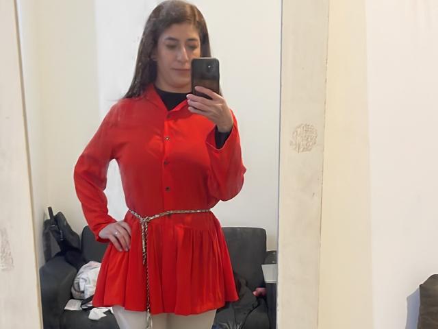 RobertWood with long brown hair, wearing a red button-up shirt and matching red skirt with white tights, standing in front of a mirror, holding a phone, showing a confident pose.