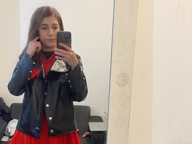RobertWood with long brown hair, wearing a black leather jacket over a red dress, holding a phone in a mirror selfie, showing a serious expression.