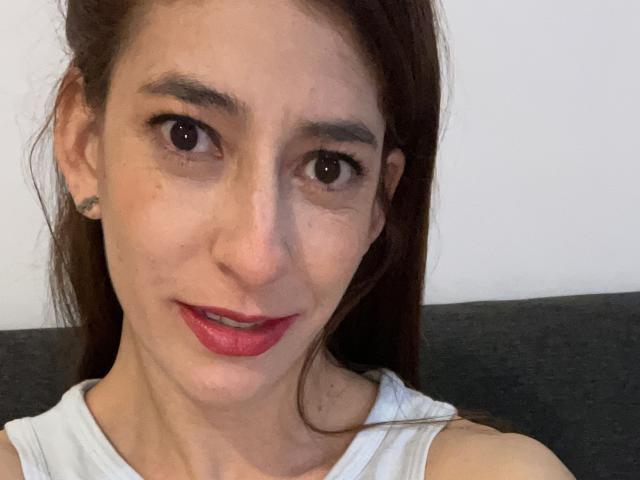 RobertWood has brown eyes, red lipstick, and a white sleeveless shirt, with brown hair and a small tattoo on the left ear, smiling slightly.