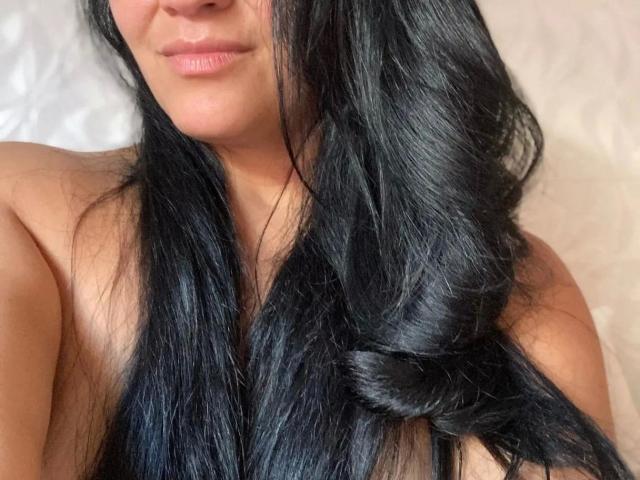 BellaGangster-hot with long, shiny black hair in a loose side braid, smiling softly, showing her cute nose and pink lips, topless, with warm skin.