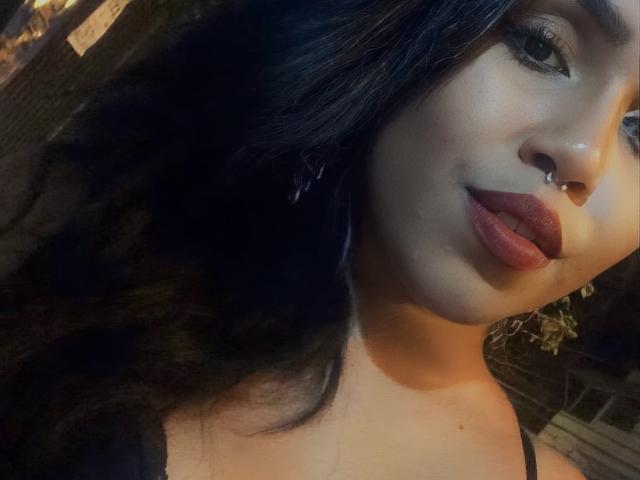 JaddieMr. has dark hair, a nose ring, and full, red lips. She's wearing a black top, looking slightly up with a soft smile. Her skin is light brown.