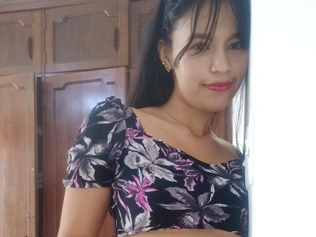 SusanKartier with medium tits, wearing a black floral crop top, smiling, has long black hair, gold earrings, and a gold necklace, standing next to wooden cabinets.