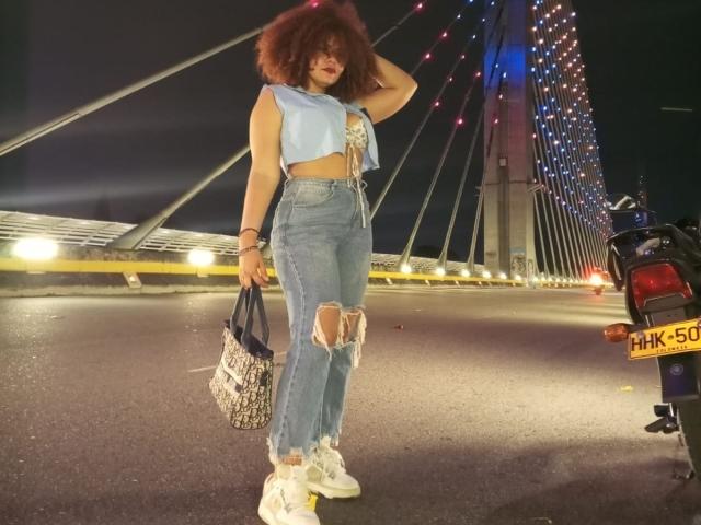 Nutellita with curly red hair, wearing a blue sleeveless crop top and ripped blue jeans, white sneakers, holding a patterned bag, standing next to a motorcycle at night.