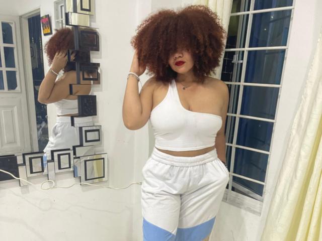Nutellita with big curly brown hair, red lipstick, white one-shoulder top, and white athletic pants with blue stripes, standing in front of a mirror, showing her ass.