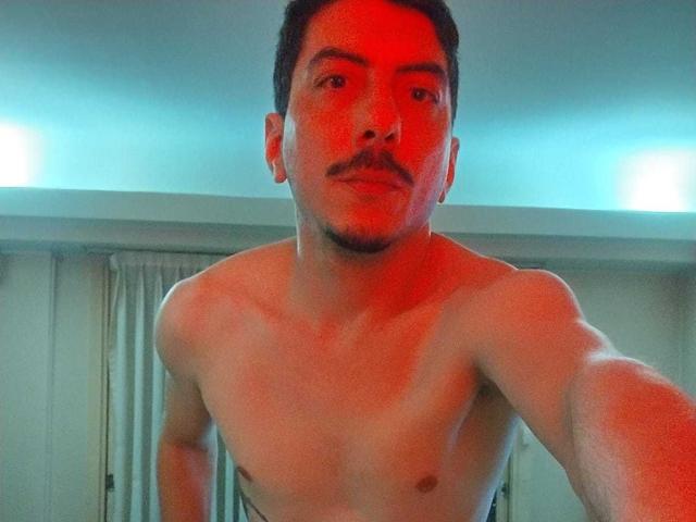 XSantyX, shirtless, red-lit, muscular, short dark hair, mustache, slight beard, taking selfie, upper body, visible chest, nipples, neutral facial expression, standing, right arm extended.