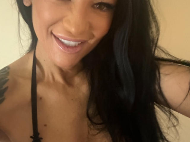 AndraD with long black hair, smiling with white teeth, showing cleavage in a black halter top, tattoo on left shoulder, brown eyes, light skin, and a cute face.