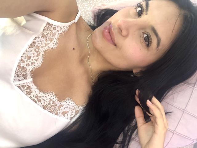 EmilyJensenn with long black hair, wearing a white lace top, smiling at the camera, showing her small, cute tits, and a delicate necklace, lying on a pink bed.