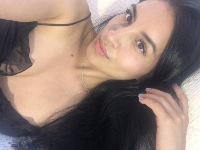 EmilyJensenn has long black hair, light skin, and is looking at the camera with pouty pink lips. She's wearing a black sheer top, showing her small tits.