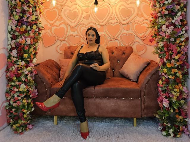 LizaRussell sits on a brown suede couch, wearing black lace top, black leather pants, and red heels, with her arms crossed, looking at the camera, surrounded by floral arrangements.