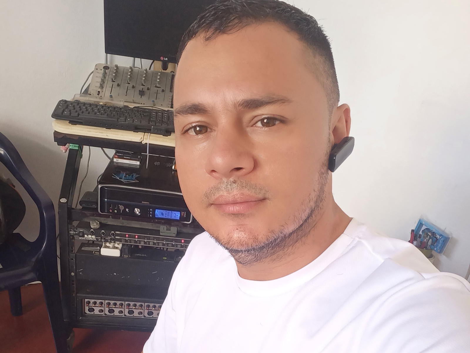 Latino69, short-haired, bearded Latino dude with medium skin, white shirt, black earbuds, serious face, sitting in front of a computer setup, looking at camera.