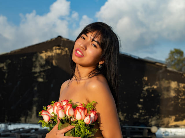 LuciaGarcia with dark hair, holding pink and white roses, bare shoulders, tan skin, and a sexy smile, against a sunlit, cloudy sky background. Boobs covered by roses.
