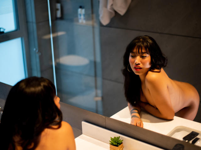LuciaGarcia, naked with dark hair, leans on bathroom sink, looking at mirror reflection, red lipstick, tattoos on arm, small potted plant, white toilet in background, grey walls.