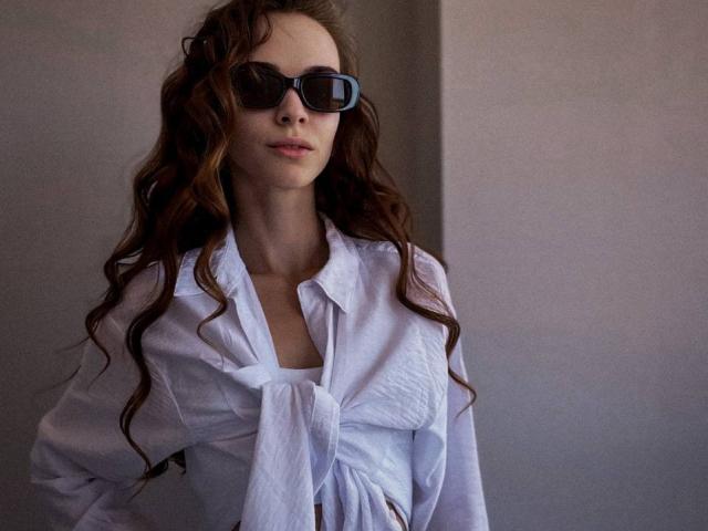 UnderTheSkin tem cabelos castanhos ondulados, usa óculos de sol escuros, uma camisa branca folgada amarrada na frente, revelando um pouco dos seios. Ela está em pé com uma expressão neutra..