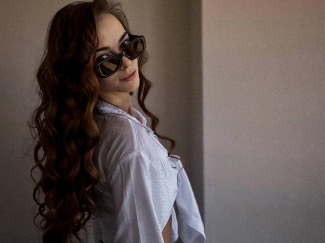 UnderTheSkin, con lunghi capelli ricci castani, che indossa occhiali da sole neri e una camicia bianca con bottoni, mostra un sorriso appena accennato, guardando oltre la spalla, con un'aria morbida, sicura e sensuale..