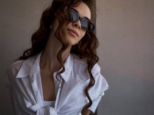 UnderTheSkin tiene el pelo castaño ondulado, lleva unas gafas negras grandes y una camisa blanca holgada, con una ligera sonrisa y la cabeza inclinada hacia un lado. Se ven los senos debajo de la camisa..
