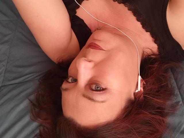 SabCoquine with red lipstick, lying on a bed, wearing black lace top, white earbuds, and red eyeshadow, looking up with pale skin and red curly hair.