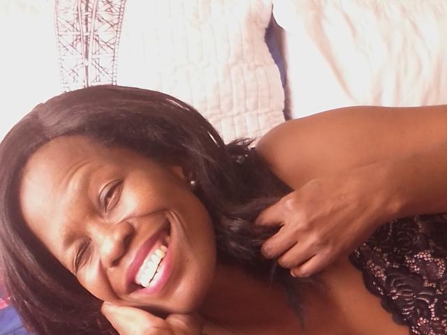 NorieHot with dark skin, smiling widely, eyes closed, black wavy hair, holding her hair with one hand, wearing a black sequined top, lying on a bed.